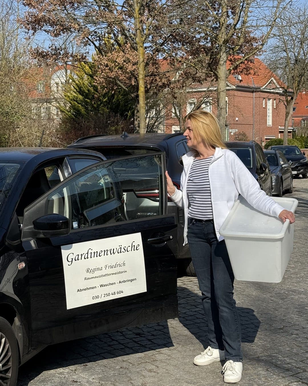 Regina Friedrich trägt eine Wäschebox zu ihrem Firmenauto mit dem Schild „Gardinenwäsche Regina Friedrich“